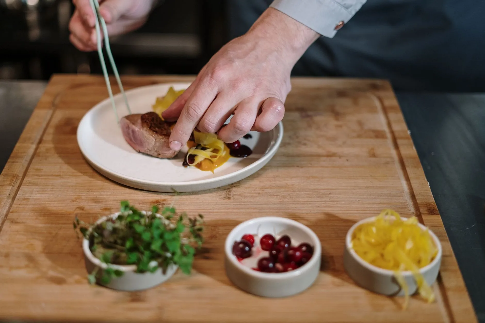 A chef's hands plating a dish on a wooden board beside small bowls of garnish