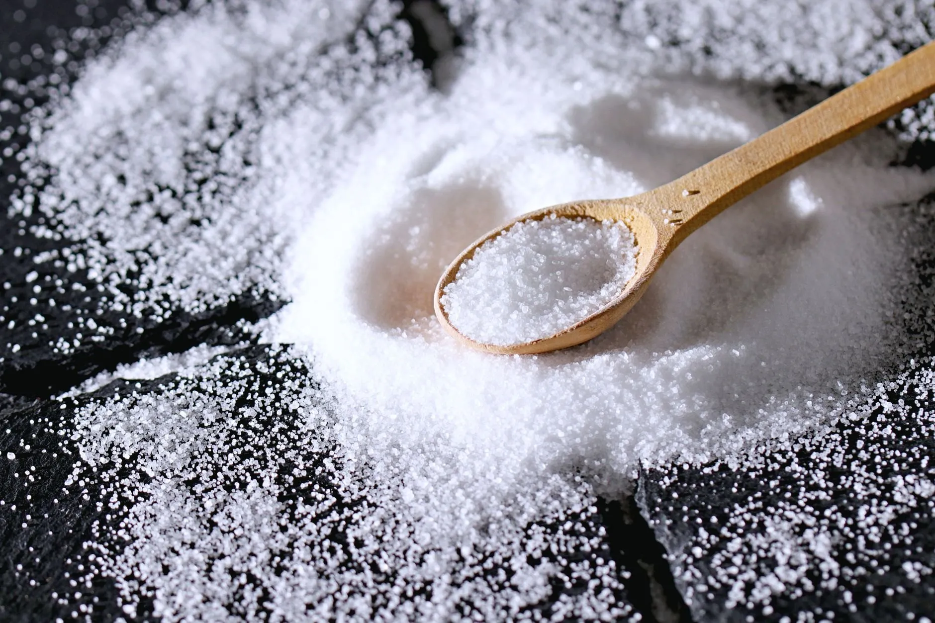 A wooden spoon full of fine white sea salt resting in a spill of salt on dark slate