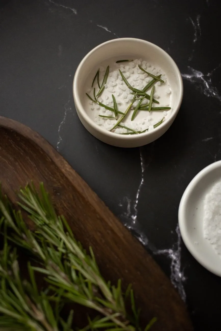 A white dish of salt flakes with chopped rosemary on dark marble