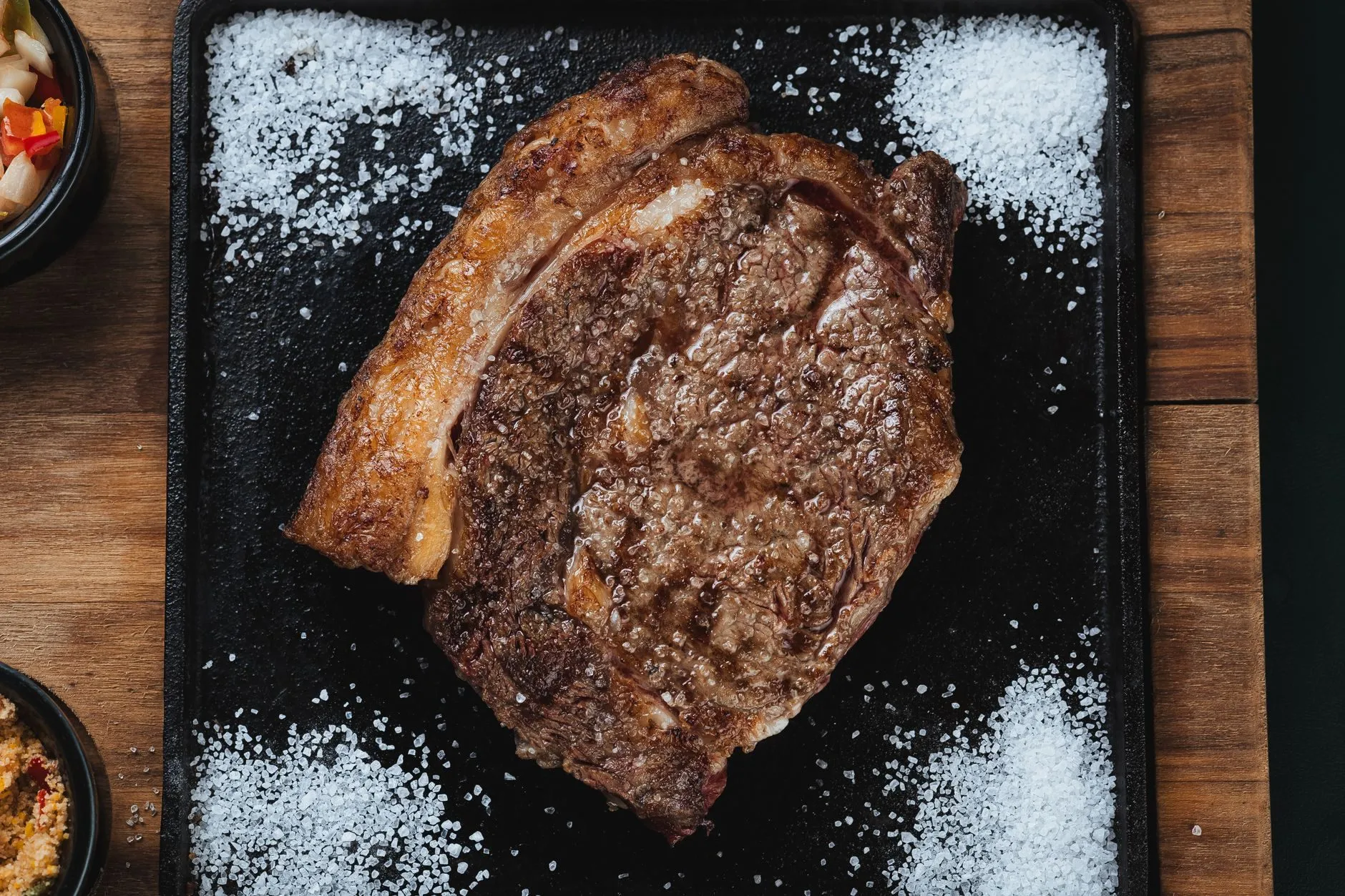 A seared rib steak on a black board with small heaps of coarse salt in each corner