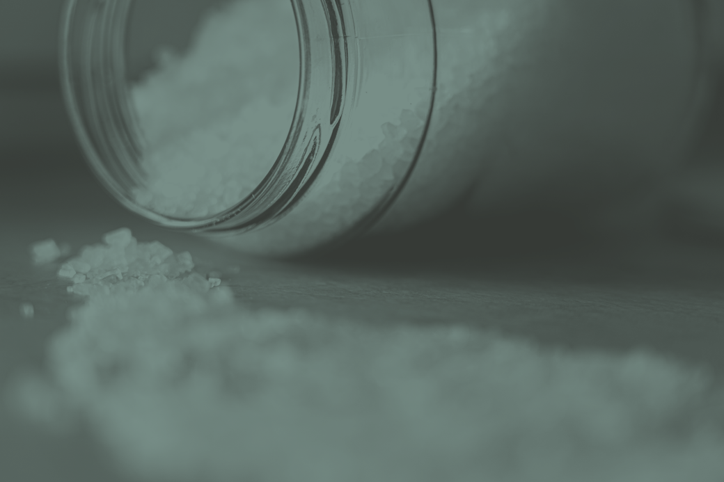 A glass kilo jar tipped on its side with coarse salt spilling out of it onto a plain surface.