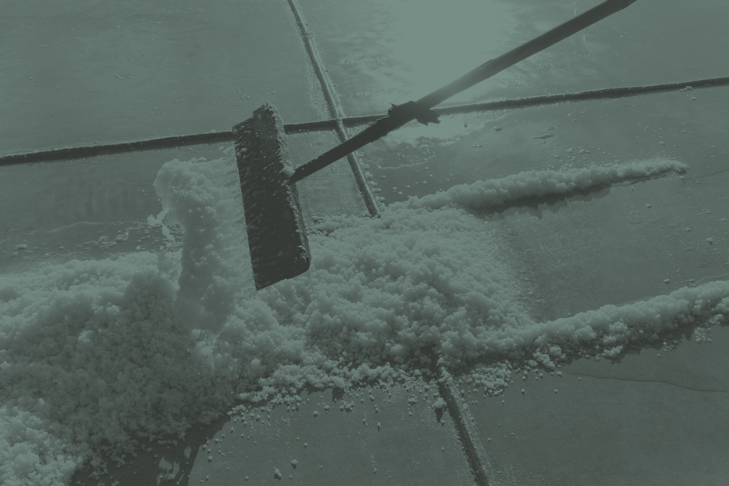 A wooden-bladed rake pushing a ridge of wet salt crystals across the flat floor of a crystalliser pan, with shallow brine standing beyond it.